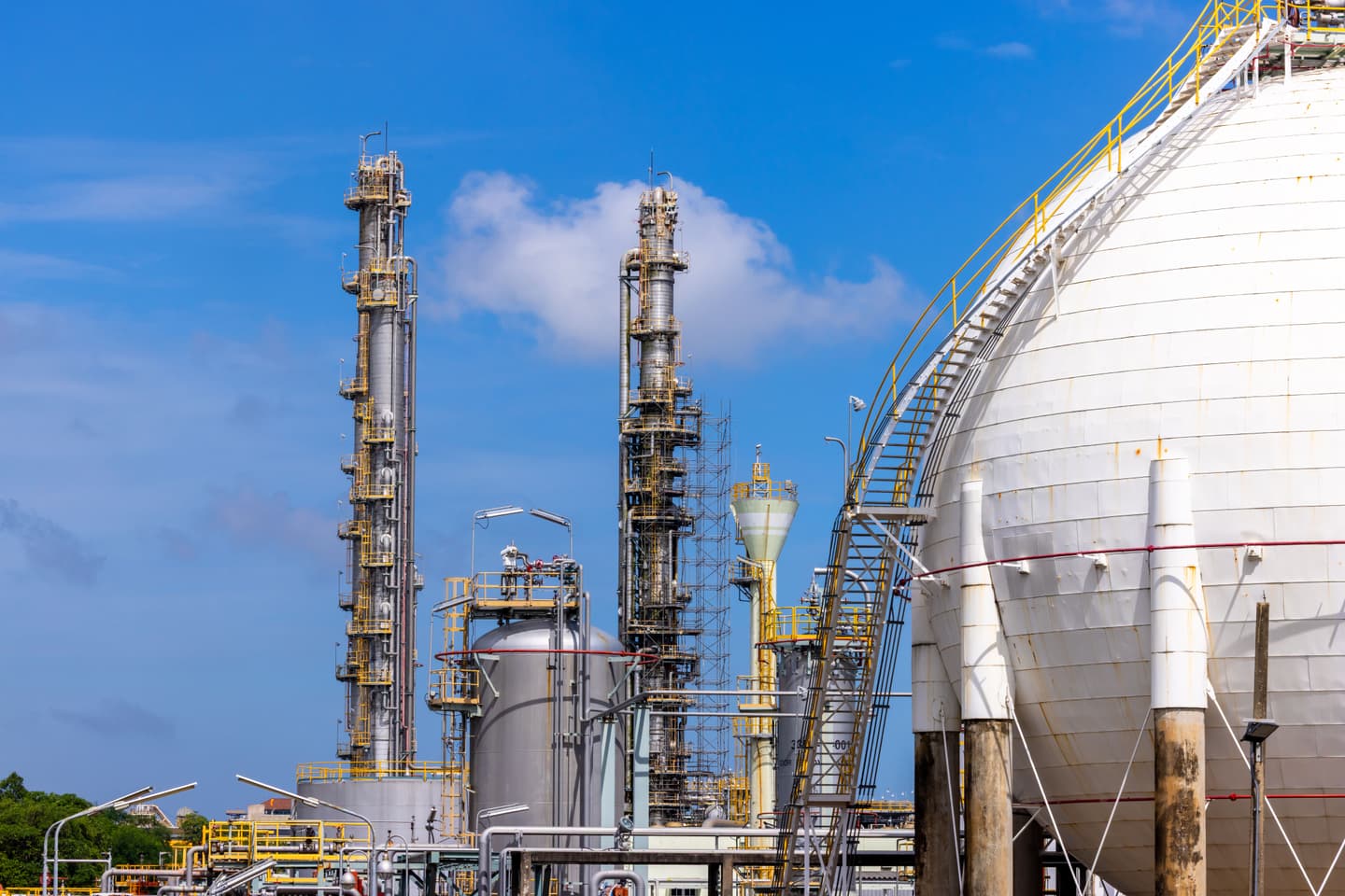 Oil refinery featuring tall industrial processing towers, interconnected pipelines and a large white spherical storage tank under a clear blue sky.