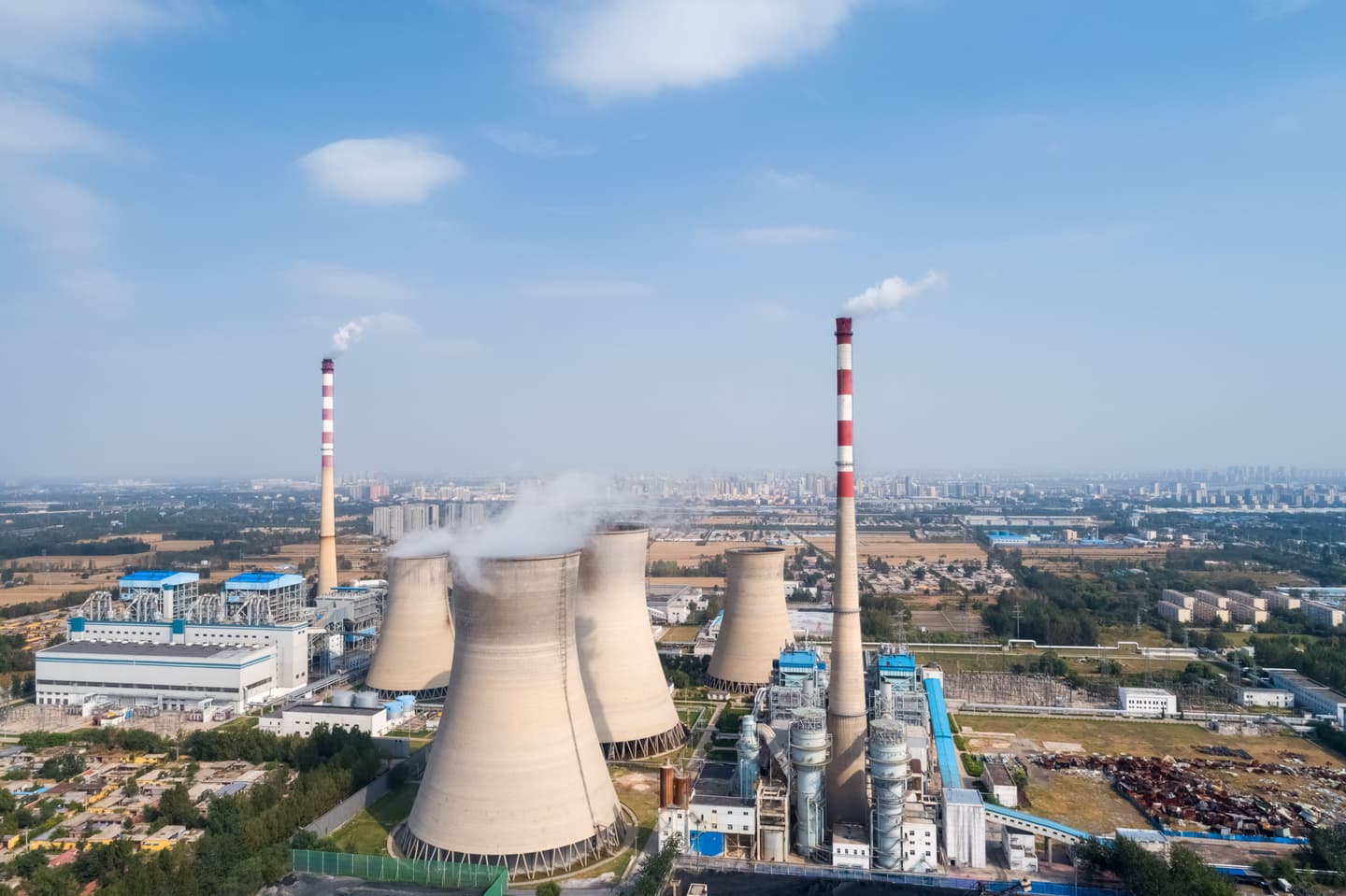 Aerial view of a thermal power plant featuring large cooling towers, industrial chimneys emitting steam, and surrounding power generation infrastructure under a clear blue sky.