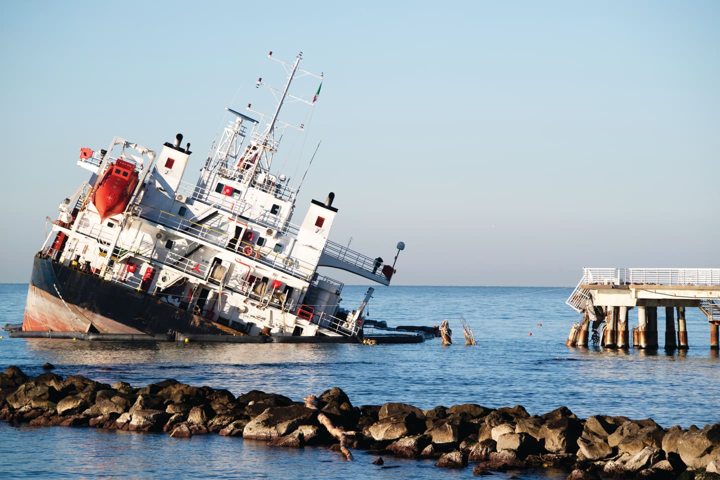 A cargo ship partially submerged and tilted near a coastal pier, illustrating maritime accidents and the challenges of marine salvage and oil spill response.