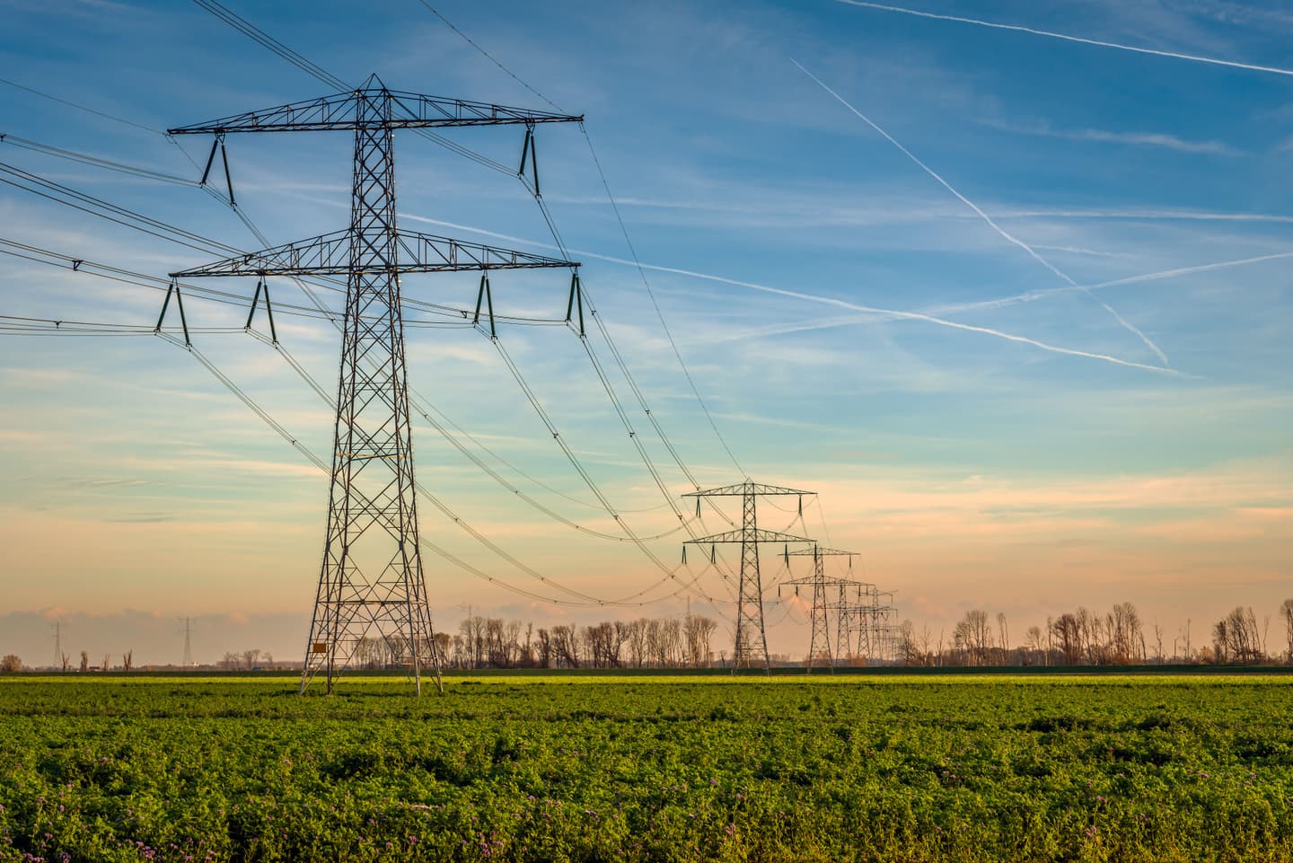High-voltage electricity transmission towers and overhead power lines stretch across a green agricultural field beneath a blue sky at sunset.