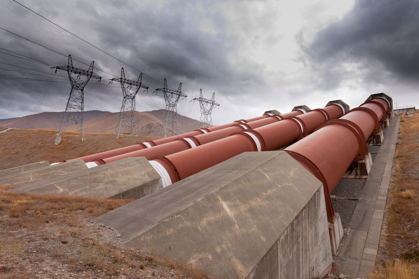Large red industrial pipelines mounted on concrete supports extend across a dry, mountainous landscape, with high-voltage electricity transmission towers and overhead power lines beneath a cloudy sky.