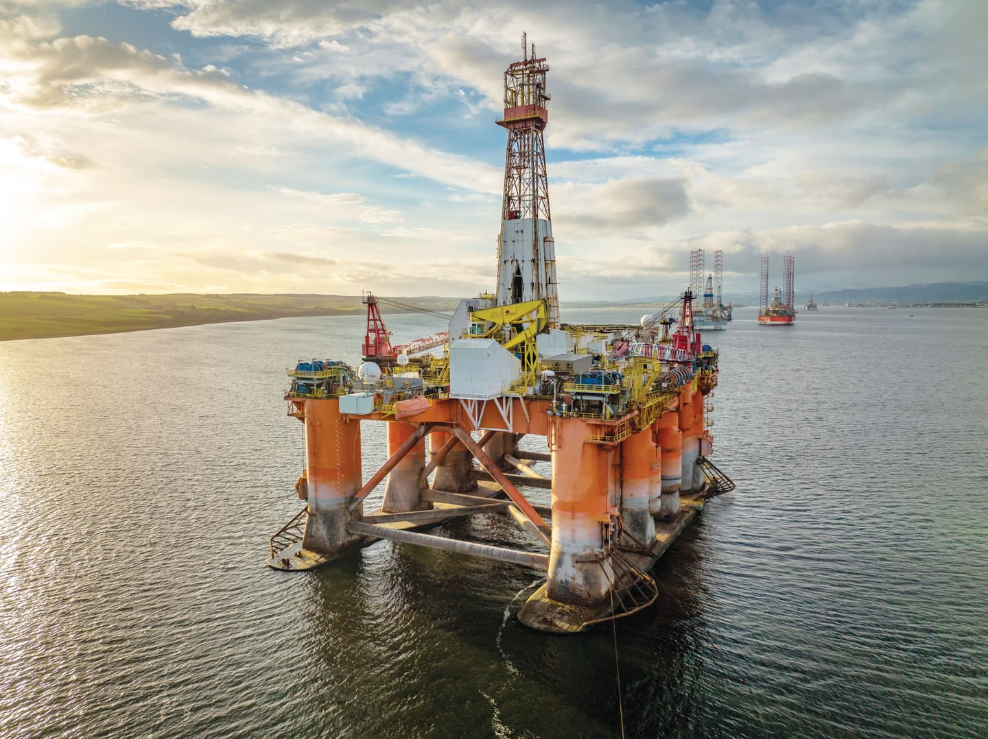 An offshore oil and gas platform standing in the sea at sunset, featuring drilling equipment, a tall derrick and supporting structures surrounded by water.