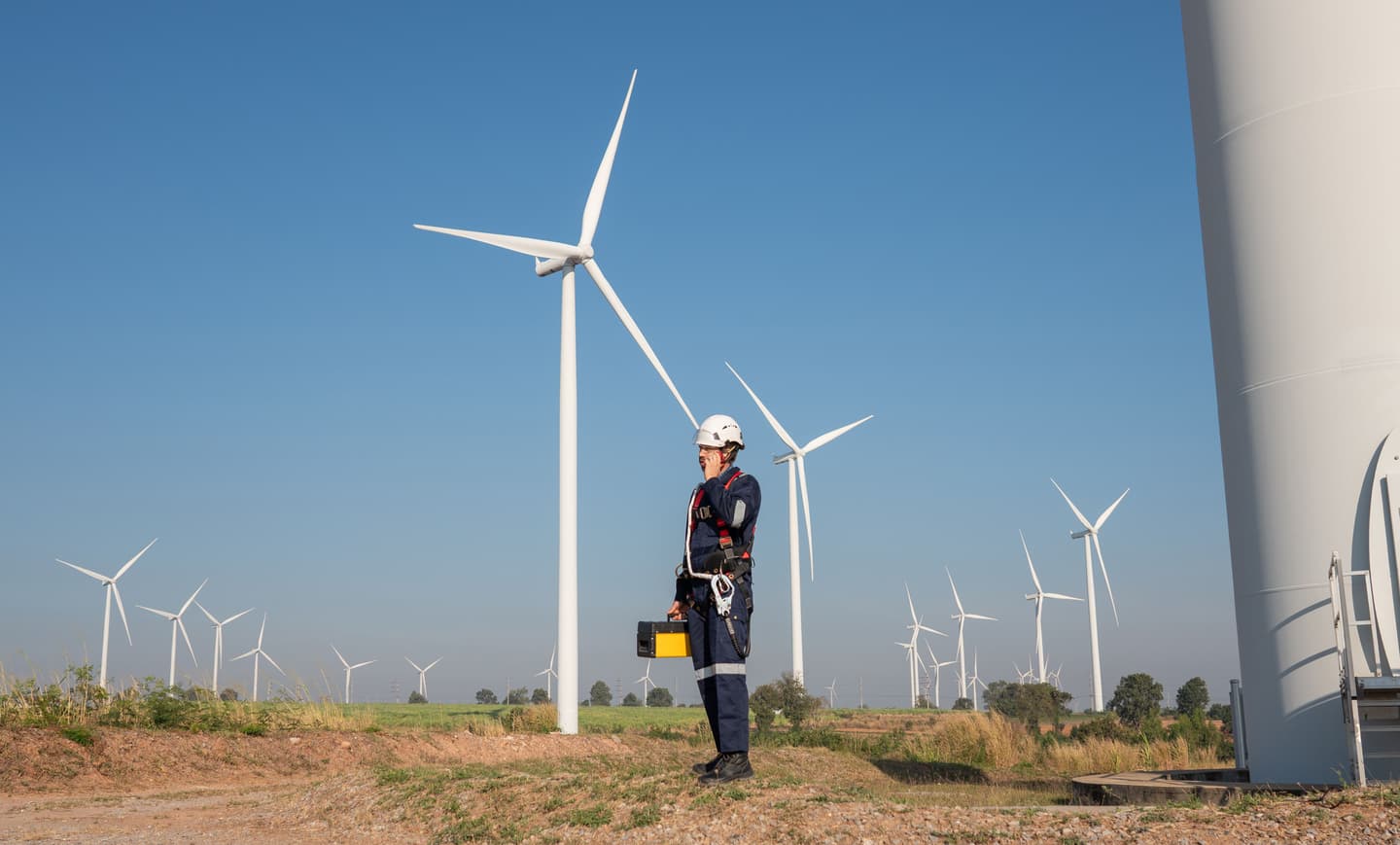 A wind turbine technician wearing a safety helmet, protective workwear and a safety harness stands beside a wind turbine at a wind farm, with multiple turbines stretching across the landscape under a clear blue sky.