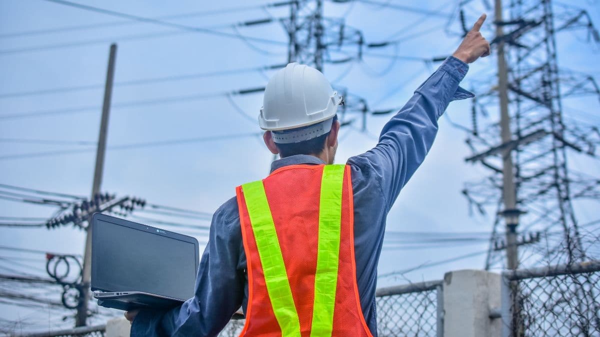Power engineer in safety gear inspecting high-voltage transmission towers with a laptop.